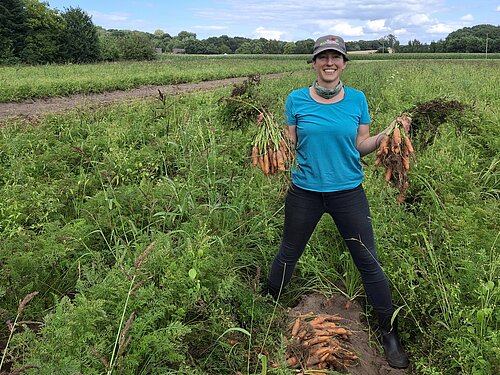 Frau auf dem Feld hält in jeder Hand ein frisch geerntetes Bund Möhren.