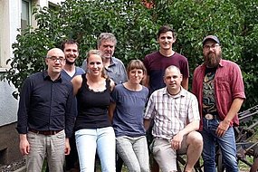 Gruppenbild mit sechs Männern und zwei Frauen vor grünem Strauch.